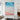 Vertical pink sand beach canvas wall art displayed next to a fireplace and grey armchair in a light-filled modern interior.